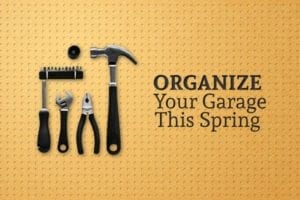 A hammer, pliers, a wrench, and a screwdriver with a variety of bits hang upon a wall beside the words "Organize Your Garage This Spring"