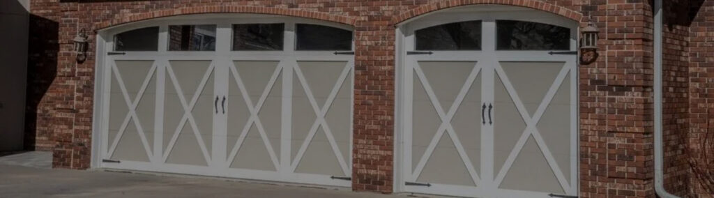 Two arched carriage-style garage doors with X-pattern wood trim and decorative handles, set into a red brick home facade with matching wall sconces on either side