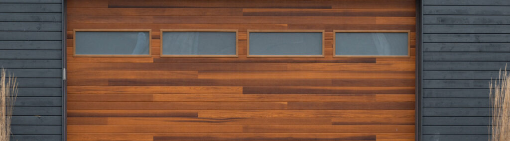 Modern dark wood garage door with horizontal planks, featuring four narrow rectangular glass windows aligned horizontally across the top section, flanked by dark grey siding.