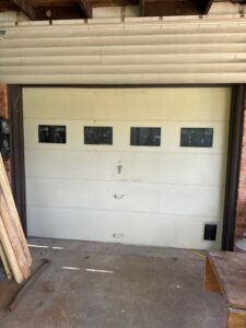 An aged, off-white sectional garage door with four small windows, central lift handles, and a pet door installed in the lower corner.