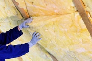 Gloved hands installing yellow fiberglass batt insulation into an attic ceiling between wooden joists.