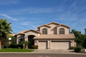 A two-story beige Mediterranean-style home with a clay tile roof, palm trees, and two beige garage doors—one single-car and one double-wide.